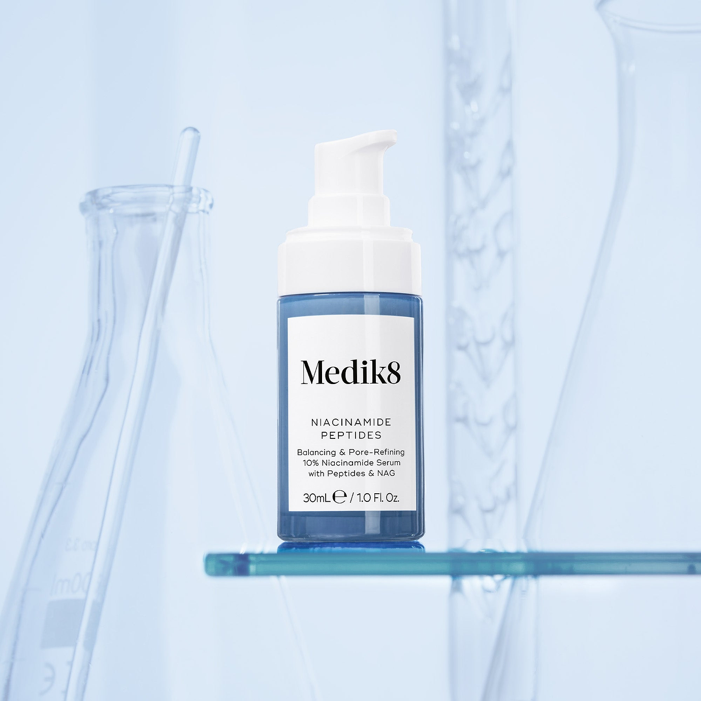 A bottle of Medik8 Niacinamide Peptides serum displayed on a glass shelf with laboratory beakers in the background. Text on the bottle highlights 