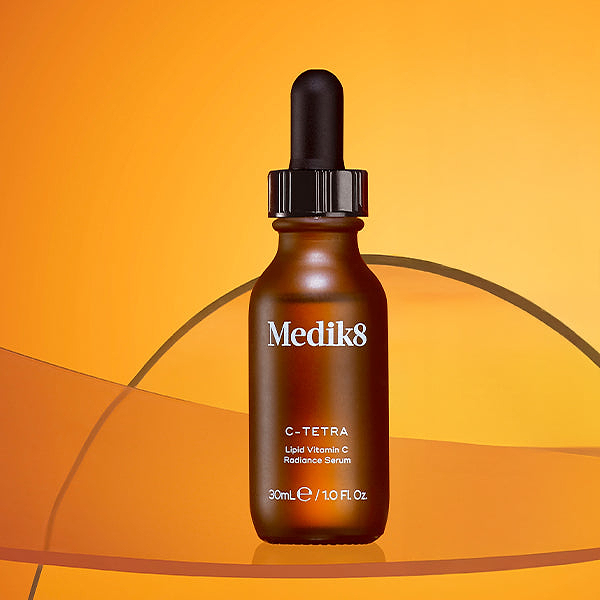 A Medik8 C-Tetra Lipid Vitamin C Radiance Serum dropper bottle displayed against a warm orange gradient background. A translucent, sweeping curved orange graphic element crosses behind and beneath the glass bottle.