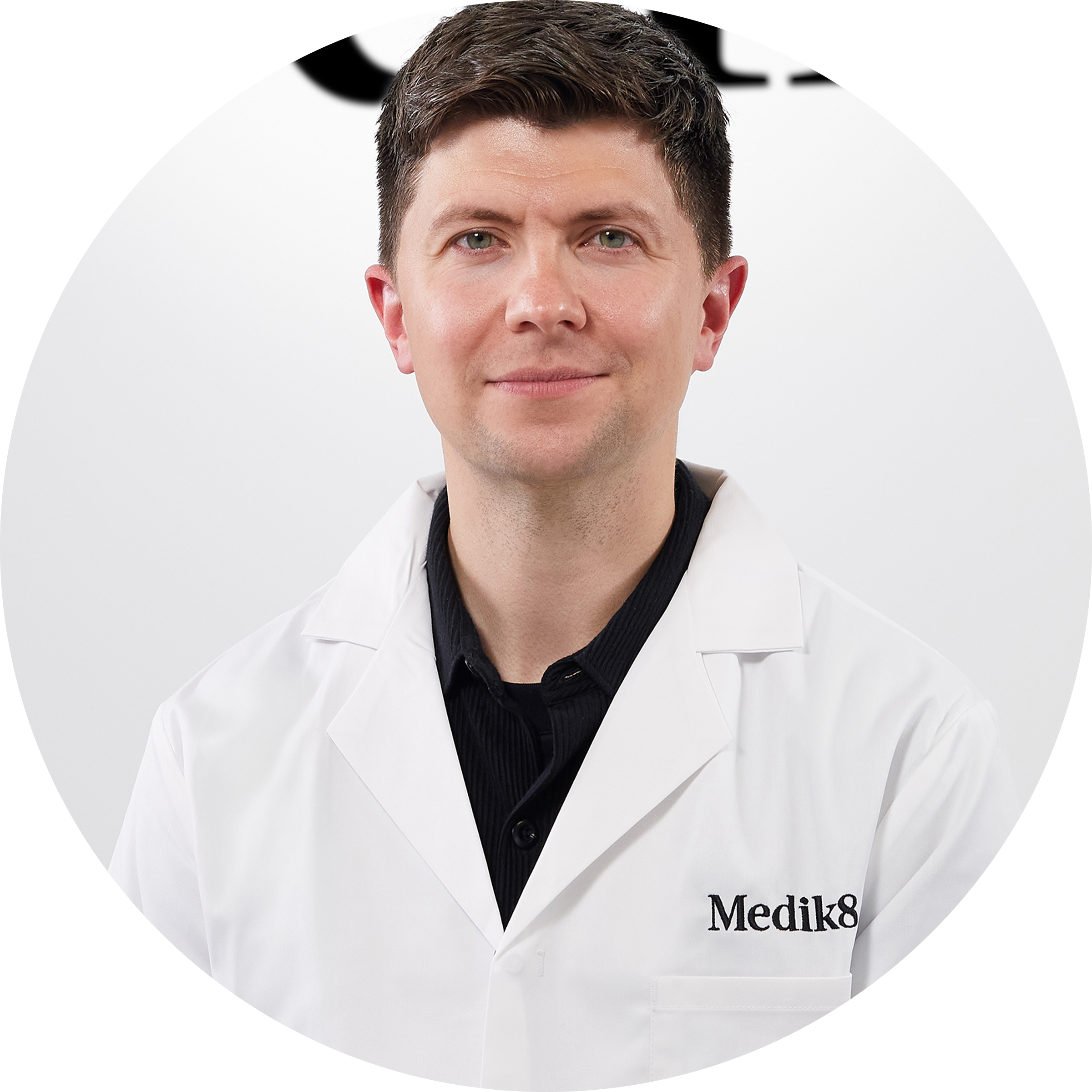A circular headshot portrait of a man with light brown hair and green eyes, smiling neutrally. He is wearing a white lab coat with 'Medik8' embroidered on the pocket and a black button-up shirt. The background is white.