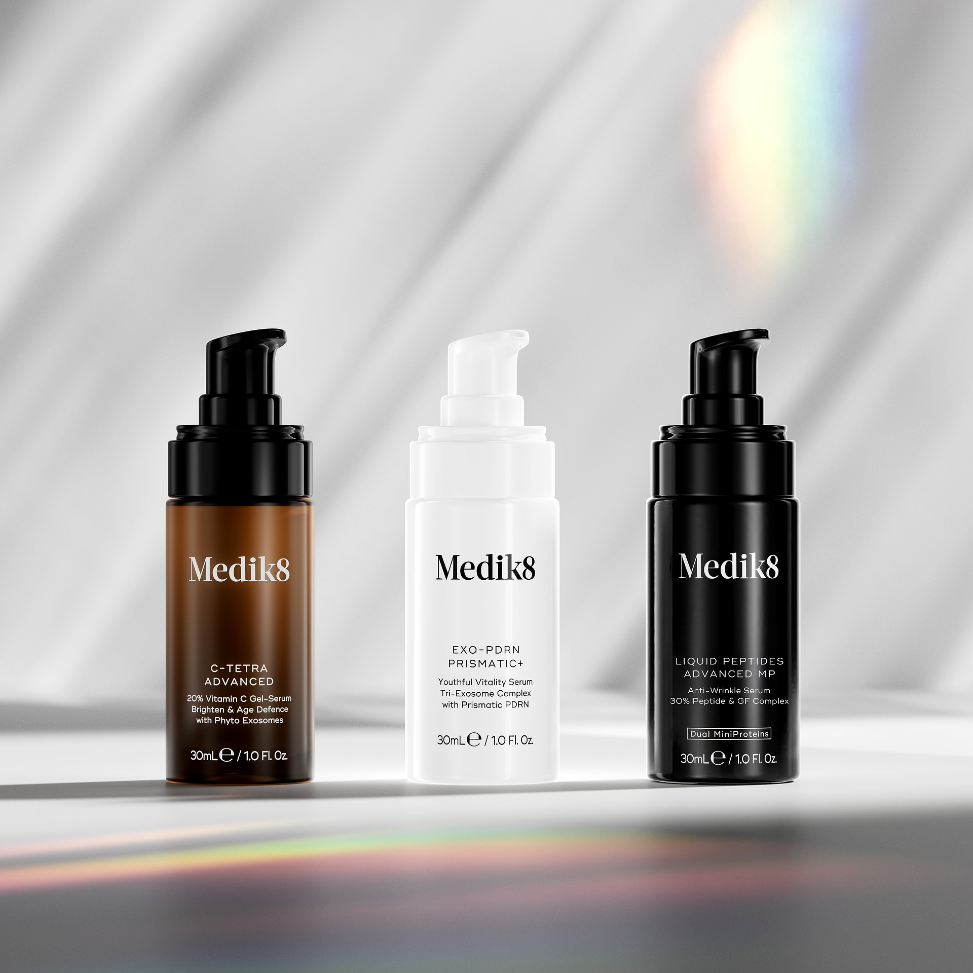 Three Medik8 pump bottles displayed on a white surface with a colourful light prism cast in the foreground and soft diagonal grey shadows in the background. From left: brown C-Tetra Advanced, white Exo-PDRN Prismatic+, and black Liquid Peptides Advanced MP. A faint rainbow light flare is visible in the top right corner.