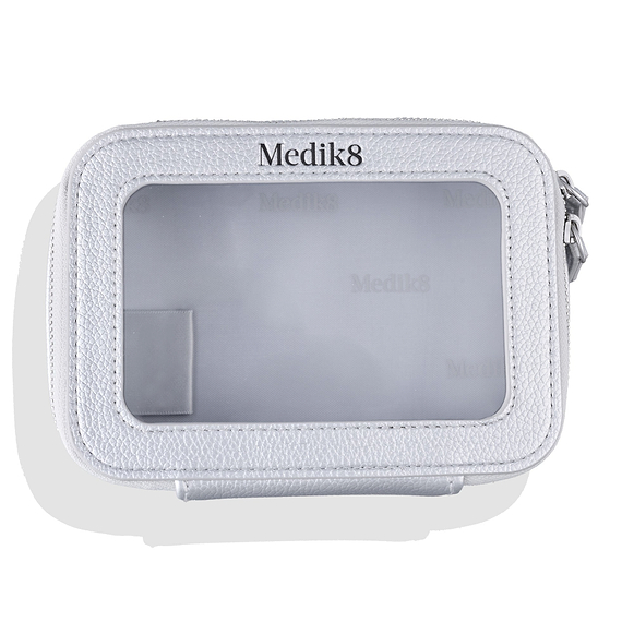 A silver faux-leather Medik8 cosmetic bag viewed from the front against a white background with a soft drop shadow. The bag features a clear, transparent window taking up most of the front panel, surrounded by a stitched border. The Medik8 logo is printed in black above the clear window.-63