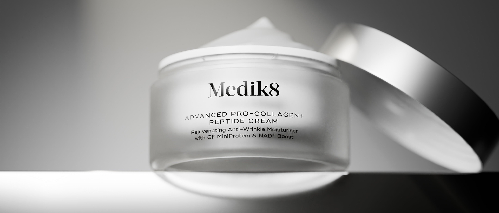 A black-and-white image of an open frosted glass jar of Medik8 Advanced Pro-Collagen+ Peptide Cream showing a peak of cream on top, with its silver lid resting on its edge behind it. The jar sits on a reflective surface against a dark grey gradient background.