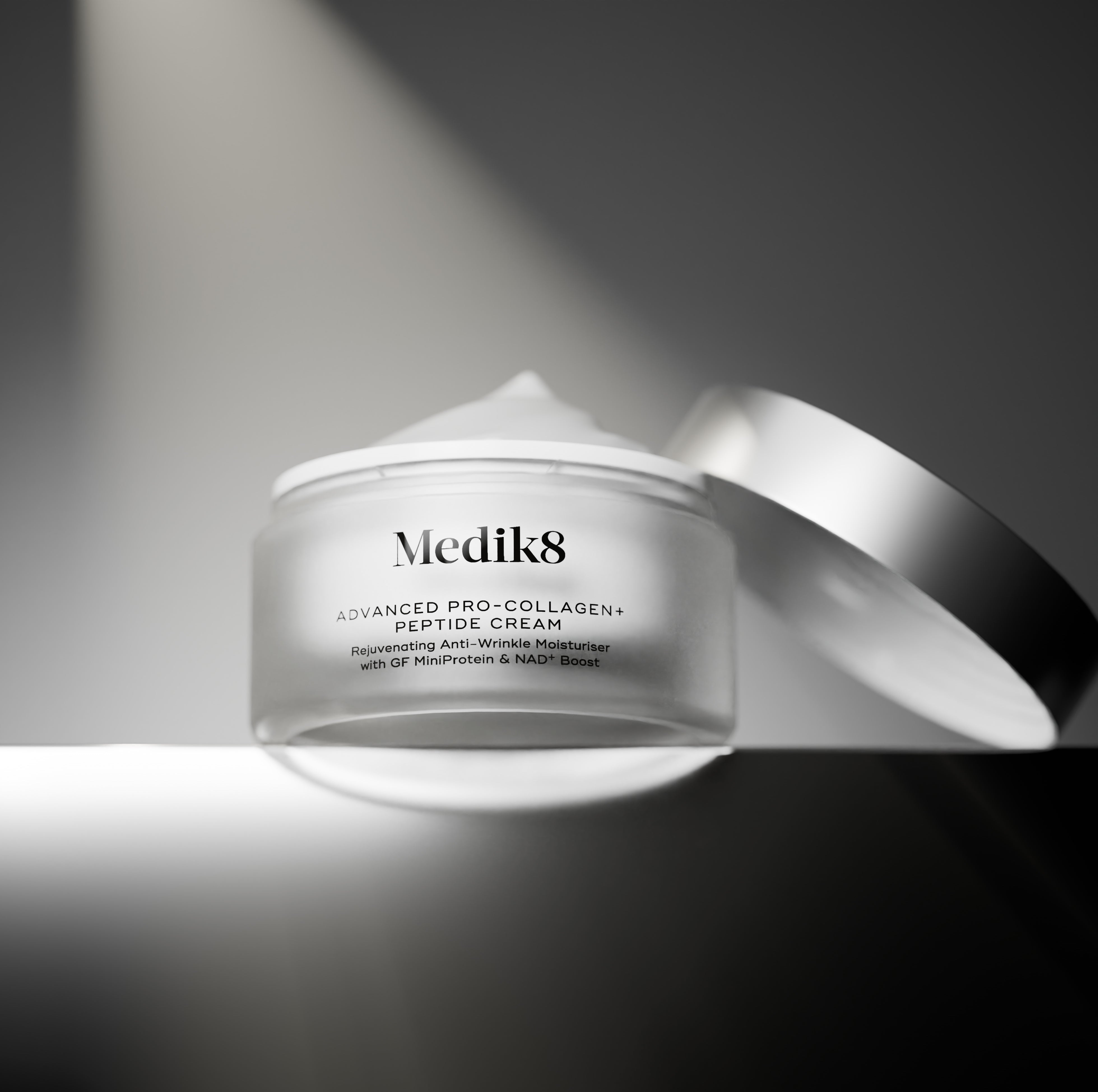 A black-and-white image of an open frosted glass jar of Medik8 Advanced Pro-Collagen+ Peptide Cream showing a peak of cream on top, with its silver lid resting on its edge behind it. The jar sits on a reflective surface against a dark grey gradient background.
