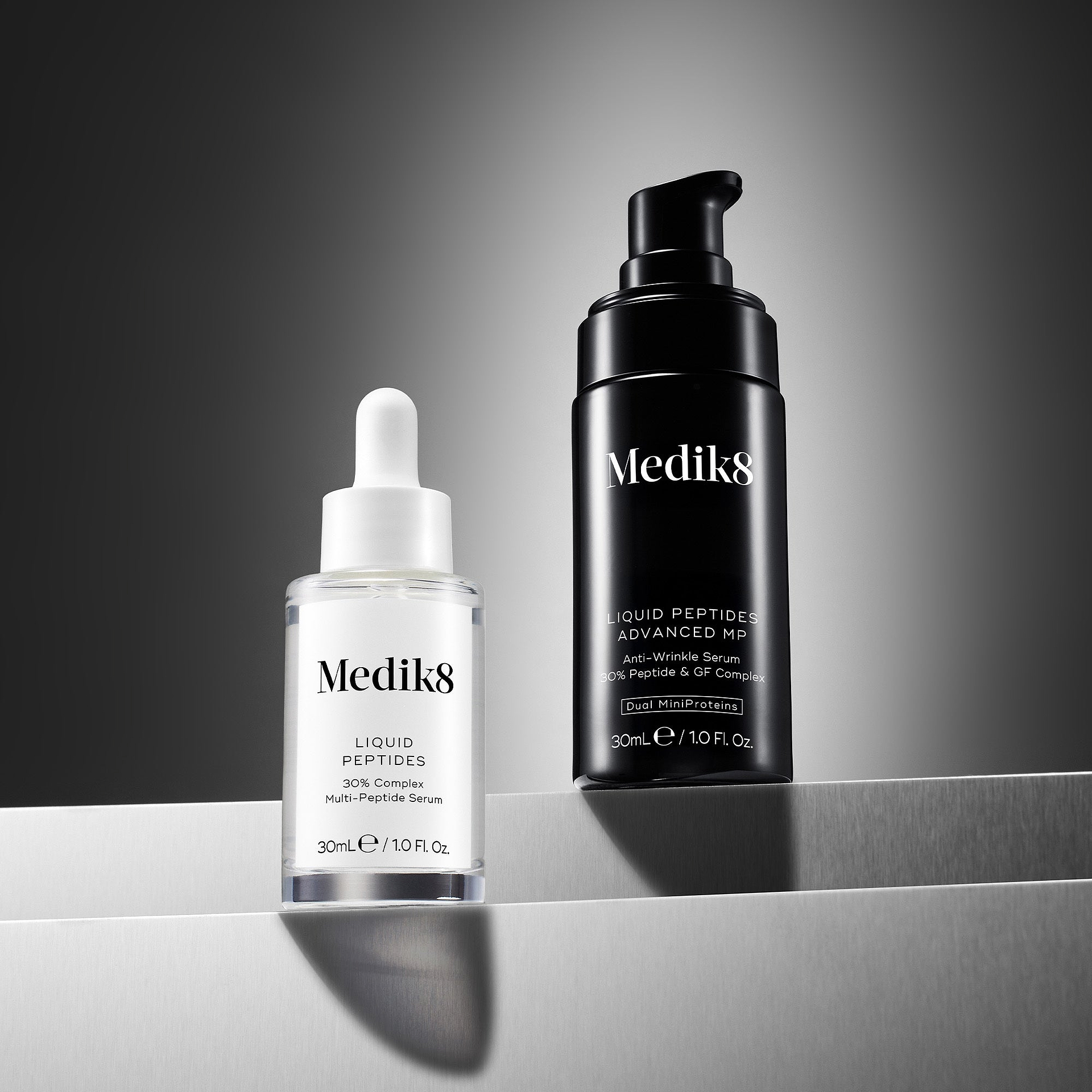 A clear Medik8 Liquid Peptides dropper bottle and a black Liquid Peptides Advanced MP pump bottle displayed on silver metallic steps against a dark grey gradient background. The clear bottle text notes a 