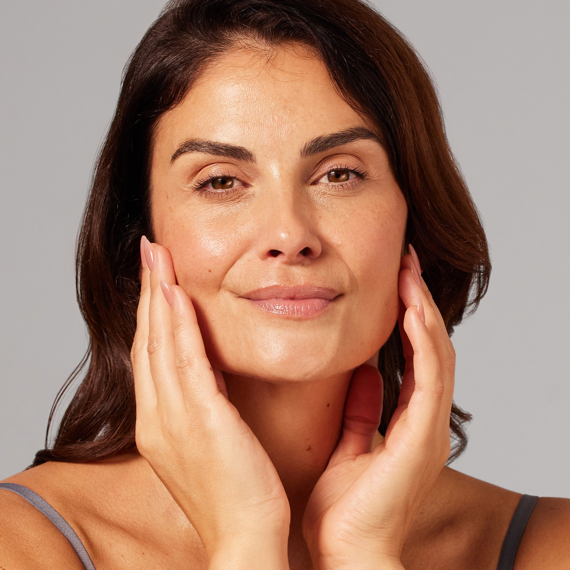 A model touches her cheeks with both hands, showcasing a radiant complexion. This close-up shot demonstrates the application of skincare products on a clean, light background.