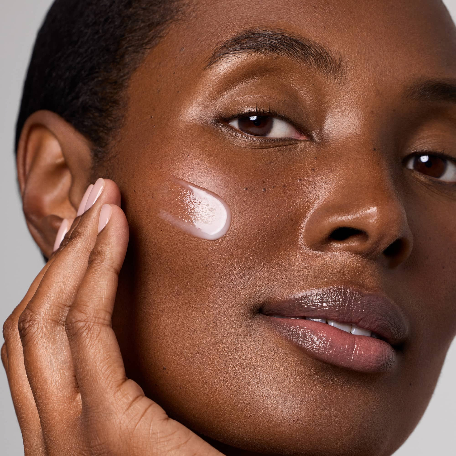 A model applies a swipe of Medik8 Liquid Peptides Advanced serum to her cheek, demonstrating its smooth, translucent texture against a neutral grey background.