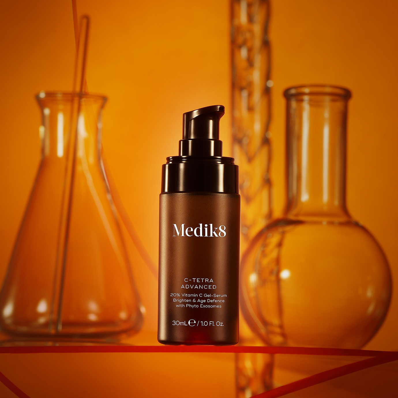 A product photo of a Medik8 C-Tetra Advanced serum bottle, staged amongst scientific laboratory flasks against a warm, orange background. The label on the brown bottle reads: 