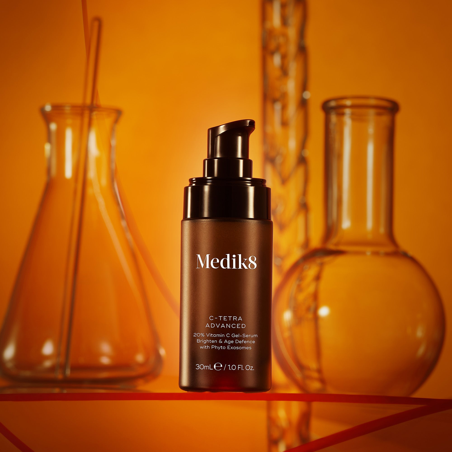 A product photo of a Medik8 C-Tetra Advanced serum bottle, staged amongst scientific laboratory flasks against a warm, orange background. The label on the brown bottle reads: 