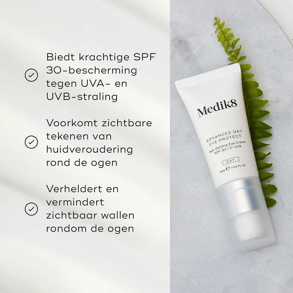 De tube Medik8 Advanced Day Eye Protect ligt op een marmeren oppervlak naast een groene varenblad. De tekst benadrukt de SPF 30-bescherming, het helpen voorkomen van zichtbare tekenen van huidveroudering en het vermogen om de oogcontour te verhelderen en wallen te verminderen.