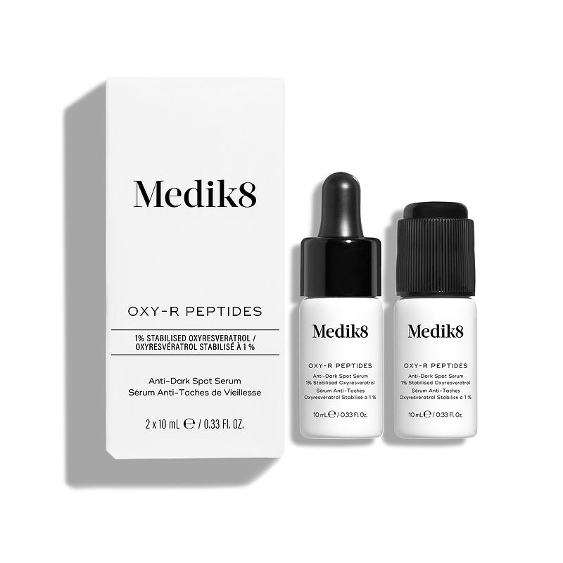 Een witte productverpakking van Medik8 Oxy-R Peptides staat naast twee kleine witte serumflacons. Eén flacon heeft een zwarte pipet, de andere een zwarte dop.