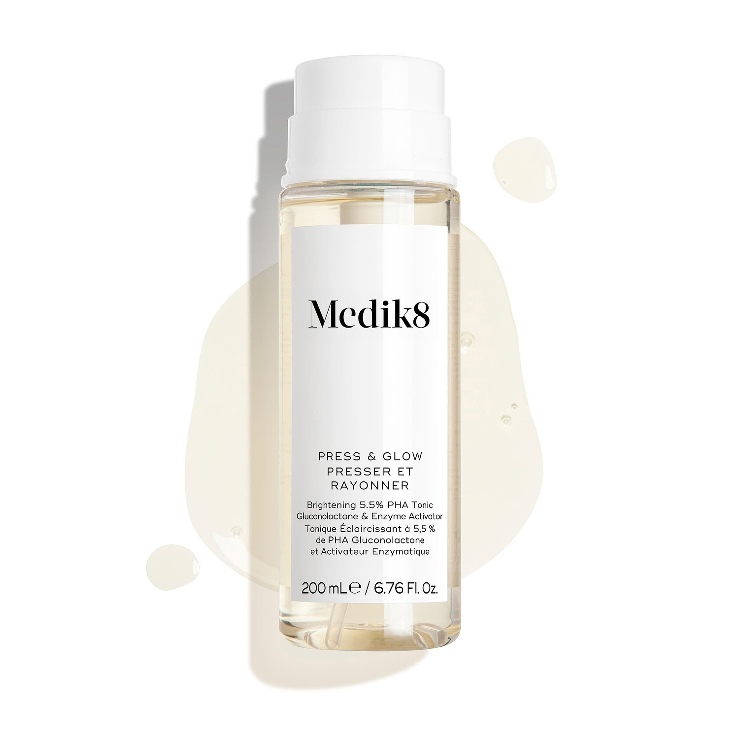 De flacon van Medik8 Press & Glow staat tegen een strakke witte achtergrond, omringd door transparante vloeistofdruppels en een klein plasje dat de waterachtige textuur benadrukt. De tekst vermeldt dat het gaat om een 200 ml Daily Exfoliating PHA Tonic with Enzyme Activator.