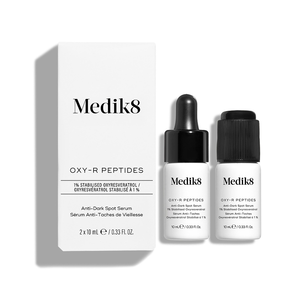 Dwie małe białe butelki serum Oxy-R Peptides są zaprezentowane na białym tle. Obok leży czarna pipeta, a na powierzchni widoczne są dwie krople żółtego serum.