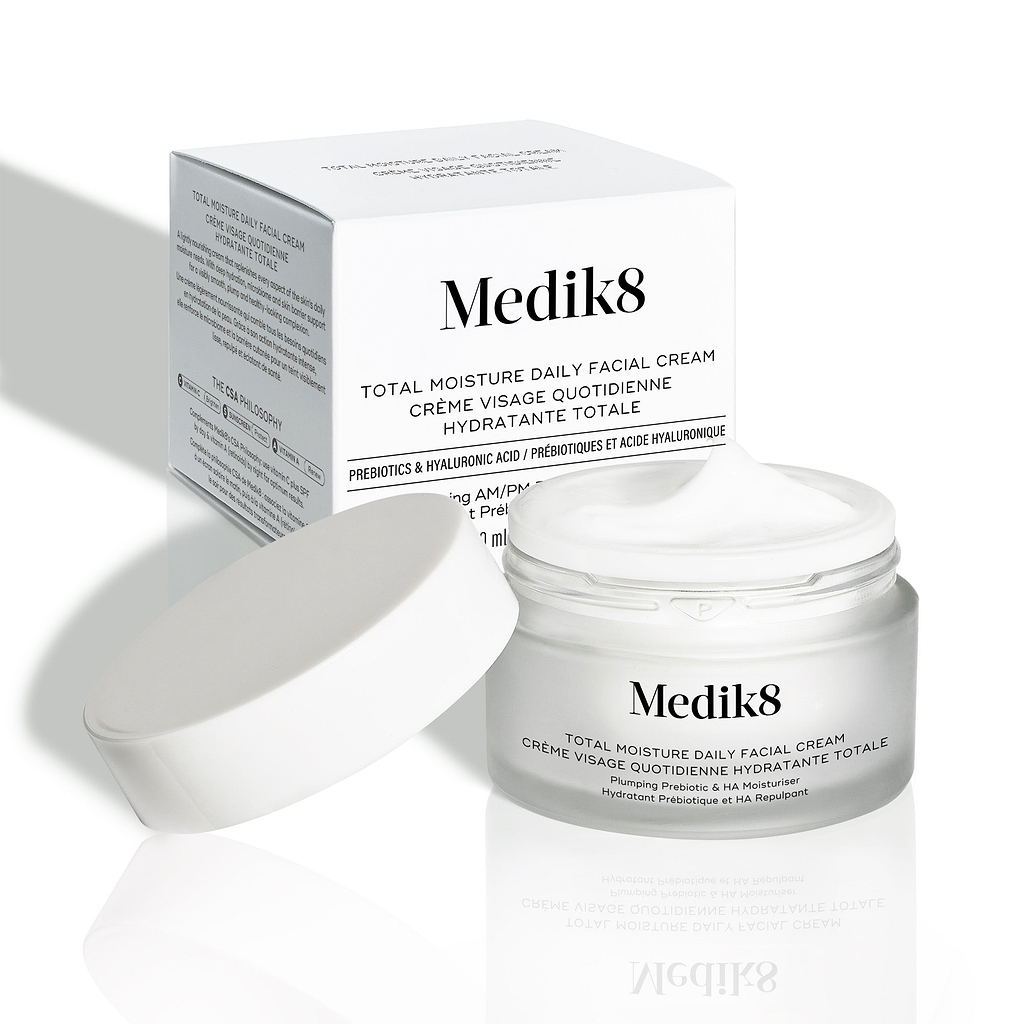 Otwarty matowy szklany słoiczek Medik8 Total Moisture Daily Facial Cream z białą zakrętką umieszczoną obok, prezentujący bogaty biały krem. Słoiczek stoi przed białym kartonowym opakowaniem na jasnym tle. Tekst na obu opakowaniach brzmi: „AM/PM wielowarstwowe nawilżenie z H.E.O.® i prebiotycznymi peptydami”.