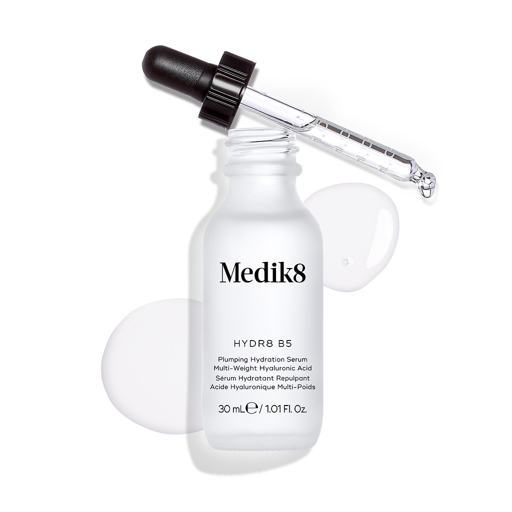 Frasco do sérum Medik8 Hydr8 B5 apresentado sobre um fundo branco limpo, com uma poça de líquido transparente e gotas à volta, com o conta-gotas apoiado no topo.