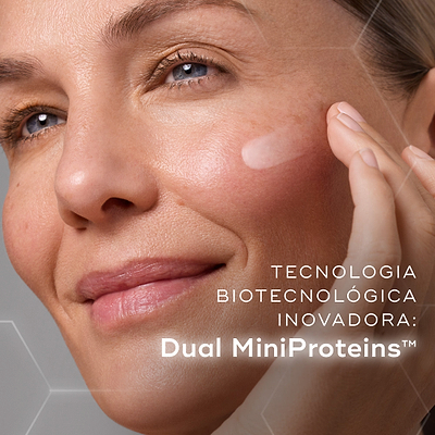 Primeiro plano de uma modella sorridente a aplicar uma porção de creme branco na bochecha contra um fundo com gráficos hexagonais subtis. O texto em português destaca a inovadora tecnologia biotecnológica: Dual MiniProteins™.