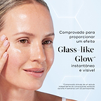 Primeiro plano de uma modella sorridente a aplicar uma porção de sérum branco na bochecha contra um fundo azul claro com gráficos hexagonais subtis. O texto em português destaca que o produto é comprovado para proporcionar um efeito Glass-like Glow instantâneo e visível.