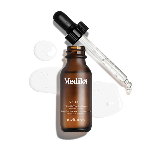 Uma imagem de produto vista de cima (“packshot”) de um frasco de 30 mL do sérum Medik8 C-Tetra  Vitamin C Serum. O frasco de vidro âmbar está sem tampa e colocado ao centro, com a tampa preta do conta-gotas apoiada na diagonal atrás do frasco. Um sérum líquido transparente está artisticamente espalhado e acumulado sobre o fundo branco limpo à volta do frasco, com algumas gotas visíveis na pipeta de vidro. A iluminação é intensa, criando sombras suaves à esquerda e uma estética clínica e elegan