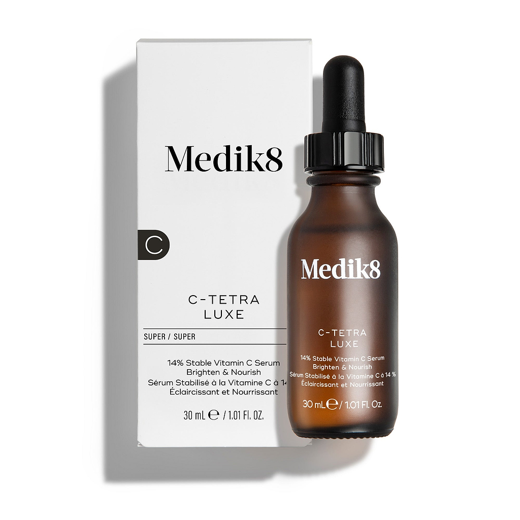 Frasco do sérum Medik8 C-Tetra Luxe e a sua embalagem de cartão branca apresentados sobre um fundo branco limpo com sombras suaves. O frasco de vidro âmbar e a caixa branca apresentam ambos o logótipo Medik8 e identificam o produto como “C-TETRA LUXE Vitamin C Serum” no formato de 30 mL.