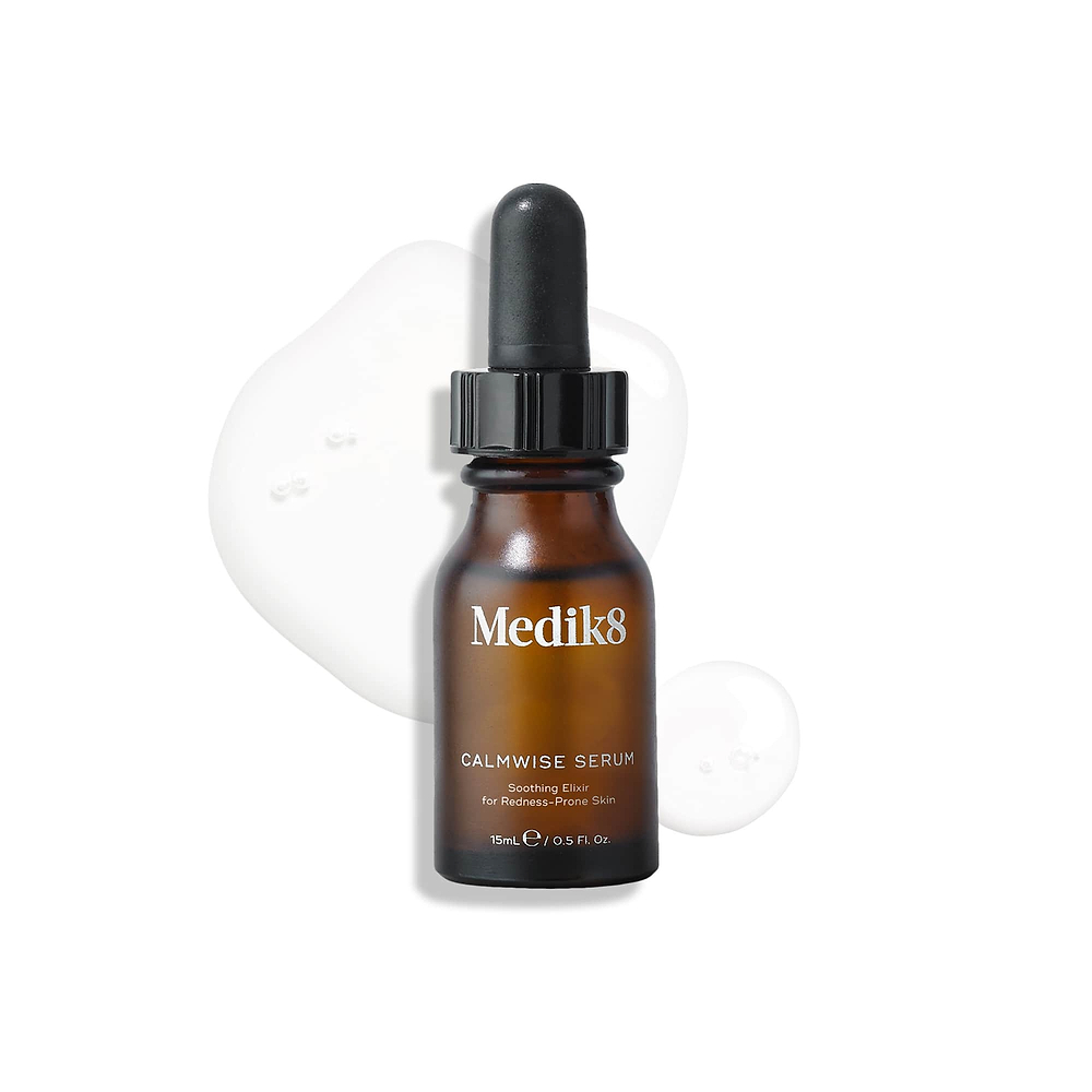 A brown glass dropper bottle of Calmwise Serum stands upright on a white surface, surrounded by clear serum droplets. 