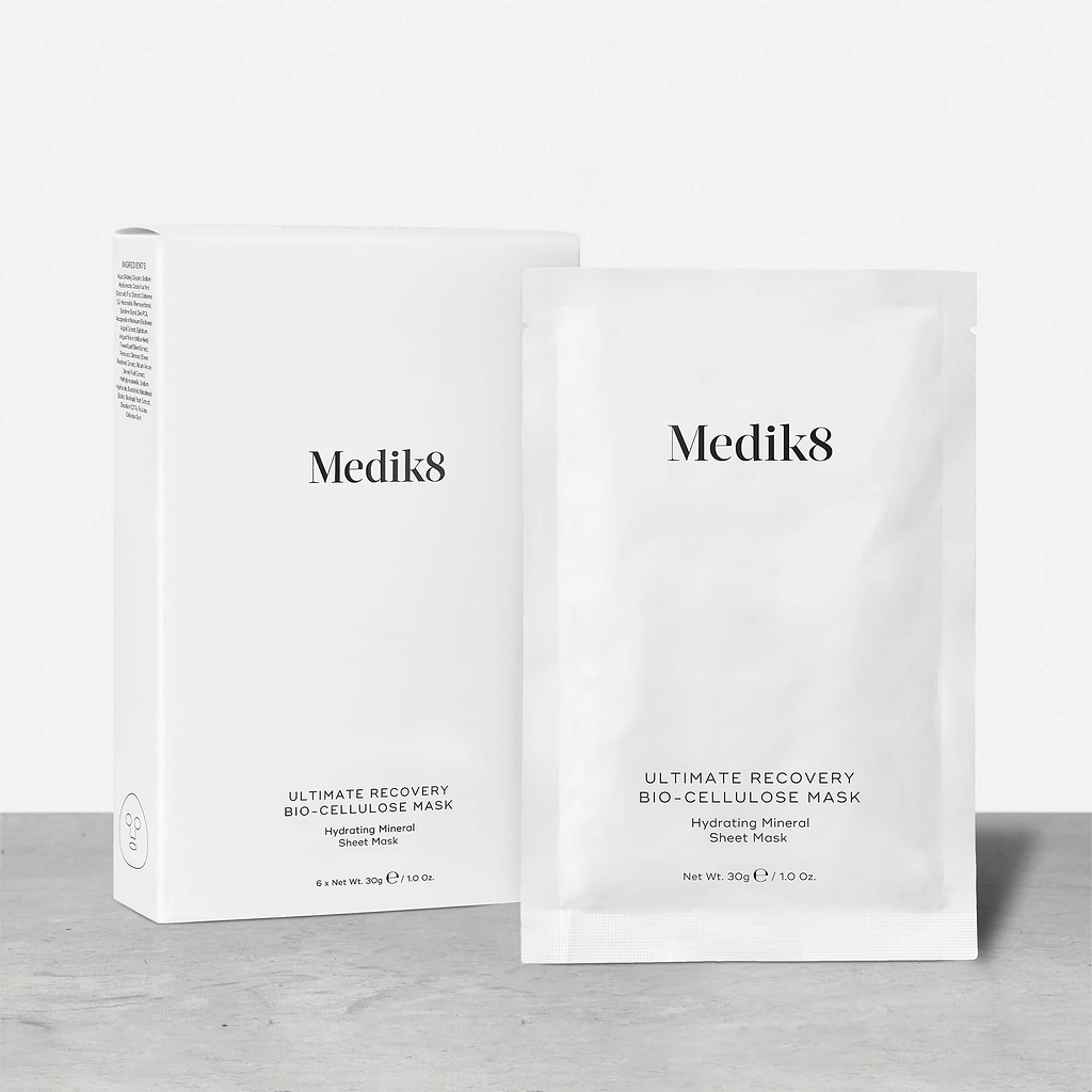 A white cardboard box of Medik8 Ultimate Recovery Bio-Cellulose Mask standing next to a single white sachet on a textured grey surface against a clean white background. Text indicates the box contains 6 sheet masks.