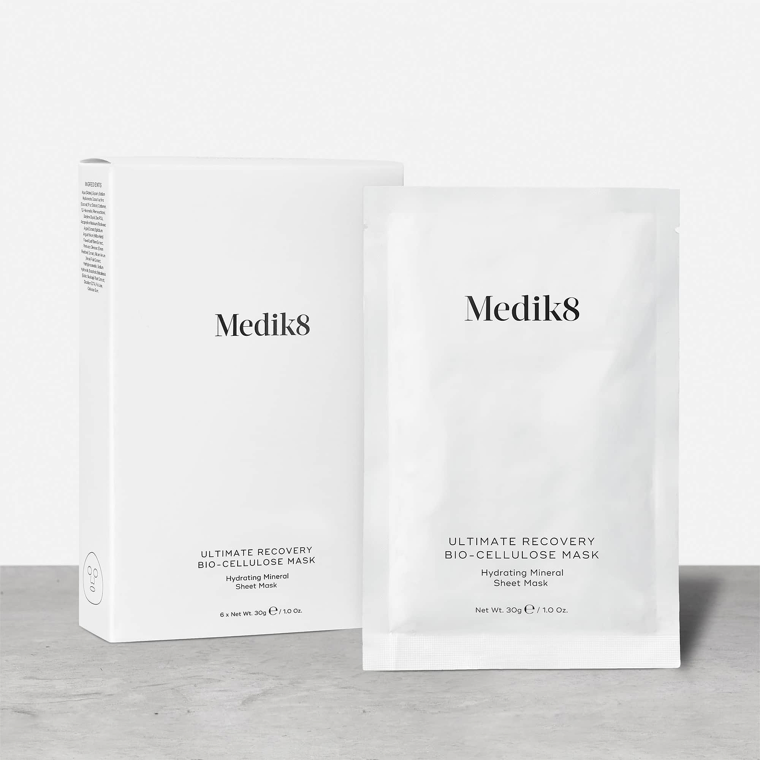 A white cardboard box of Medik8 Ultimate Recovery Bio-Cellulose Mask standing next to a single white sachet on a textured grey surface against a clean white background. Text indicates the box contains 6 sheet masks.