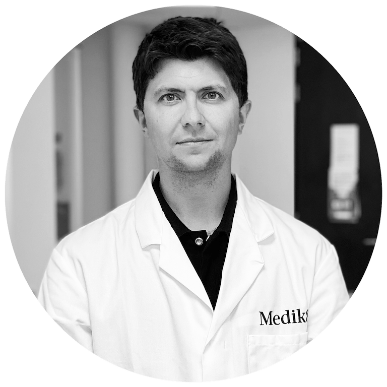 A black-and-white portrait of a Medik8 skincare scientist and founder wearing a branded lab coat, representing the research and innovation behind the formulation.
