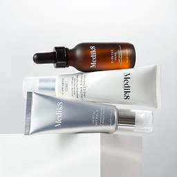 A stack of Medik8 skincare products balanced on a white block against a white background. From top to bottom: C-Tetra Luxe serum bottle (Liquid Vitamin C), Advanced Day Ultimate Protect tube (Age-Defying Moisturiser with SPF 50+), and Crystal Retinal 6 tube (Stable Retinal Night Serum).