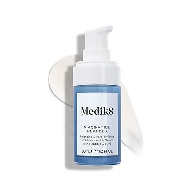 Medik8 Niacinamide Peptides serum bottle with a white pump top, resting on a white surface with a smear of white serum behind it.