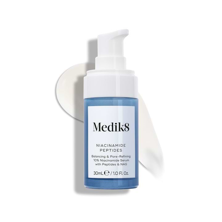 Medik8 Niacinamide Peptides serum bottle with a white pump top, resting on a white surface with a smear of white serum behind it.