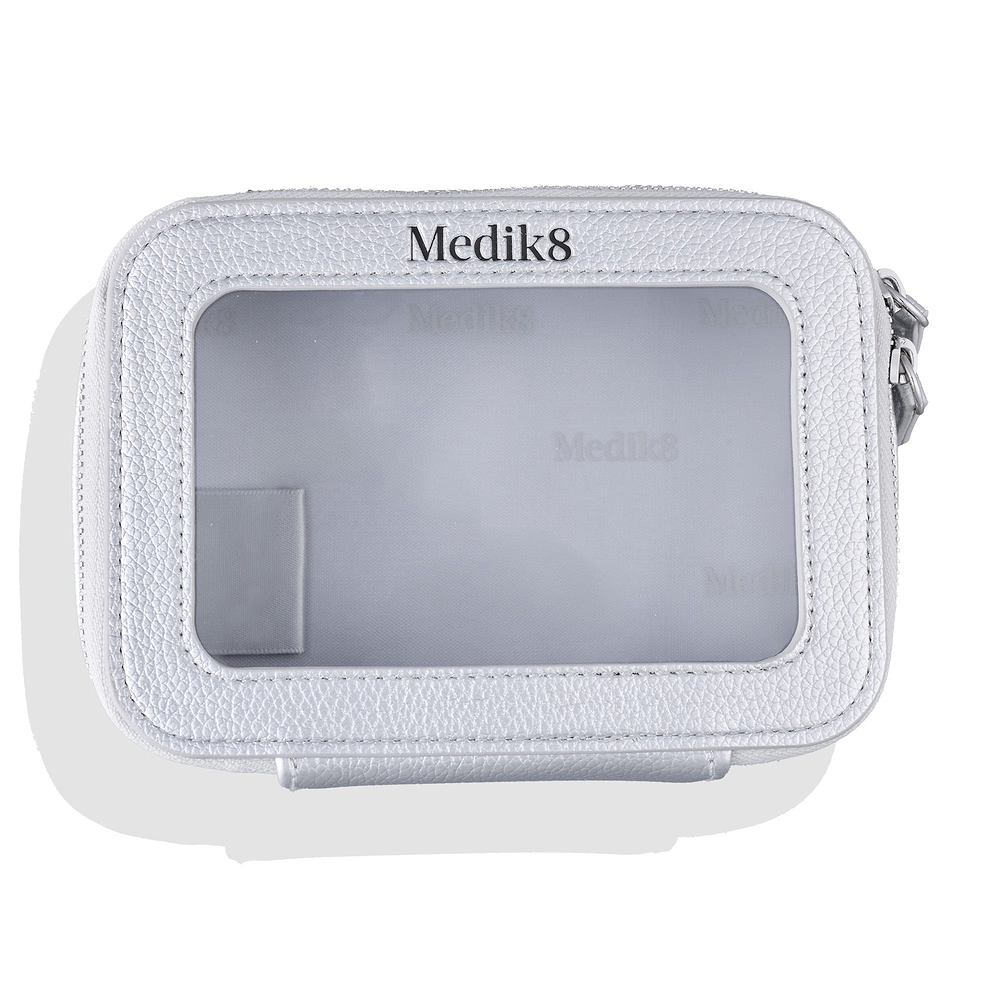 A silver faux-leather Medik8 cosmetic bag viewed from the front against a white background with a soft drop shadow. The bag features a clear, transparent window taking up most of the front panel, surrounded by a stitched border. The Medik8 logo is printed in black above the clear window.