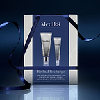 A Medik8 "Retinal Recharge" gift box wrapped with a dark blue ribbon on a reflective surface against a dark blue background. The white box features a dark blue center panel displaying the printed products inside: a Crystal Retinal 3 tube and a Crystal Retinal Ceramide Eye 3 tube. Text highlights "Age-Defying Face & Under Eye Duo."