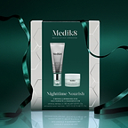 A Medik8 "Nighttime Nourish" gift box wrapped with a dark green ribbon on a reflective surface against a dark green background. The white box features a dark green center panel displaying the printed products inside: a Crystal Retinal 3 tube and an Advanced Night Restore travel size jar. Text highlights "Firming & Renewing Duo."