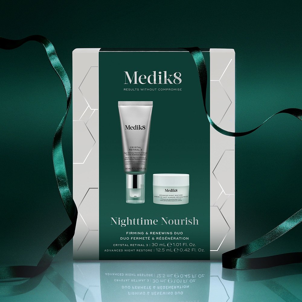 A Medik8 "Nighttime Nourish" gift box wrapped with a dark green ribbon on a reflective surface against a dark green background. The white box features a dark green center panel displaying the printed products inside: a Crystal Retinal 3 tube and an Advanced Night Restore travel size jar. Text highlights "Firming & Renewing Duo."