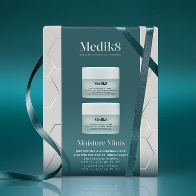 A Medik8 "Moisture Minis" gift box wrapped with a teal ribbon on a reflective surface against a teal background. The white box features a teal center panel displaying the printed products inside: a Daily Radiance Vitamin C travel size jar and an Advanced Night Restore travel size jar. Text highlights "Protecting & Nourishing Duo."