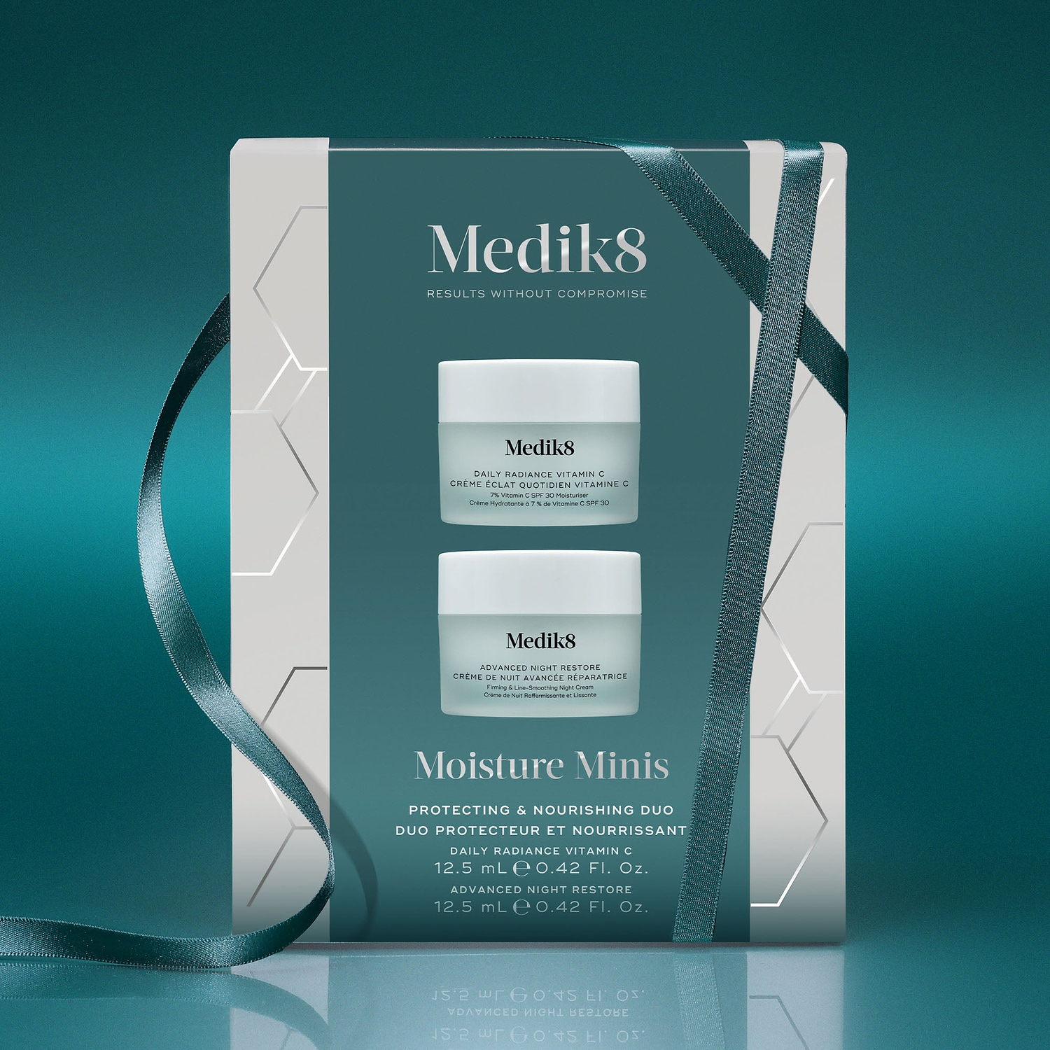 A Medik8 "Moisture Minis" gift box wrapped with a teal ribbon on a reflective surface against a teal background. The white box features a teal center panel displaying the printed products inside: a Daily Radiance Vitamin C travel size jar and an Advanced Night Restore travel size jar. Text highlights "Protecting & Nourishing Duo."