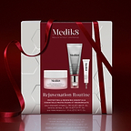 A Medik8 "Rejuvenation Routine" gift box wrapped with a red ribbon on a reflective surface against a deep red background. The white box features a deep red center panel displaying the printed products inside: a Daily Radiance Vitamin C jar, a Crystal Retinal 3 tube, and an Exo-PDRN Prismatic+ deluxe sample. Text highlights "Protecting & Renewing Essentials."
