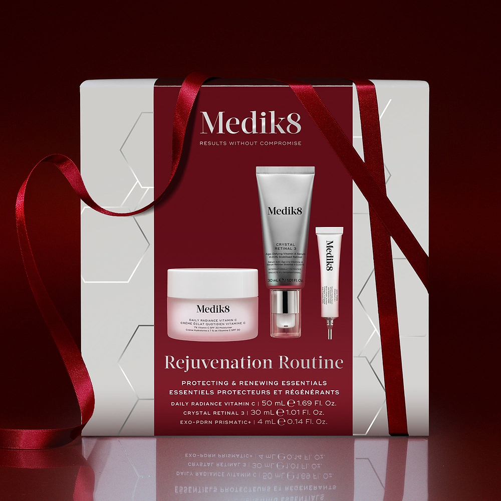 A Medik8 "Rejuvenation Routine" gift box wrapped with a red ribbon on a reflective surface against a deep red background. The white box features a deep red center panel displaying the printed products inside: a Daily Radiance Vitamin C jar, a Crystal Retinal 3 tube, and an Exo-PDRN Prismatic+ deluxe sample. Text highlights "Protecting & Renewing Essentials."