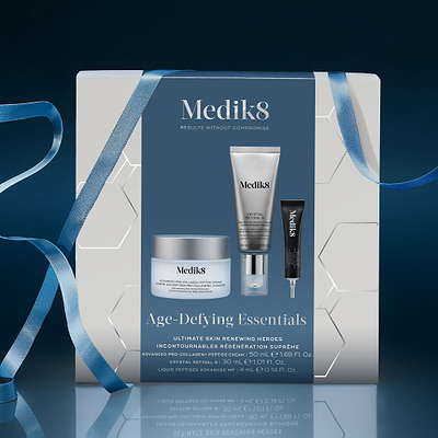 A Medik8 "Age-Defying Essentials" gift box wrapped with a blue ribbon on a reflective surface against a dark blue background. The white box features a dark blue center panel displaying the printed products inside: an Advanced Pro-Collagen+ Peptide Cream jar, a Crystal Retinal 6 tube, and a Liquid Peptides Advanced MP deluxe sample. Text highlights "Ultimate Skin Renewing Heroes."
