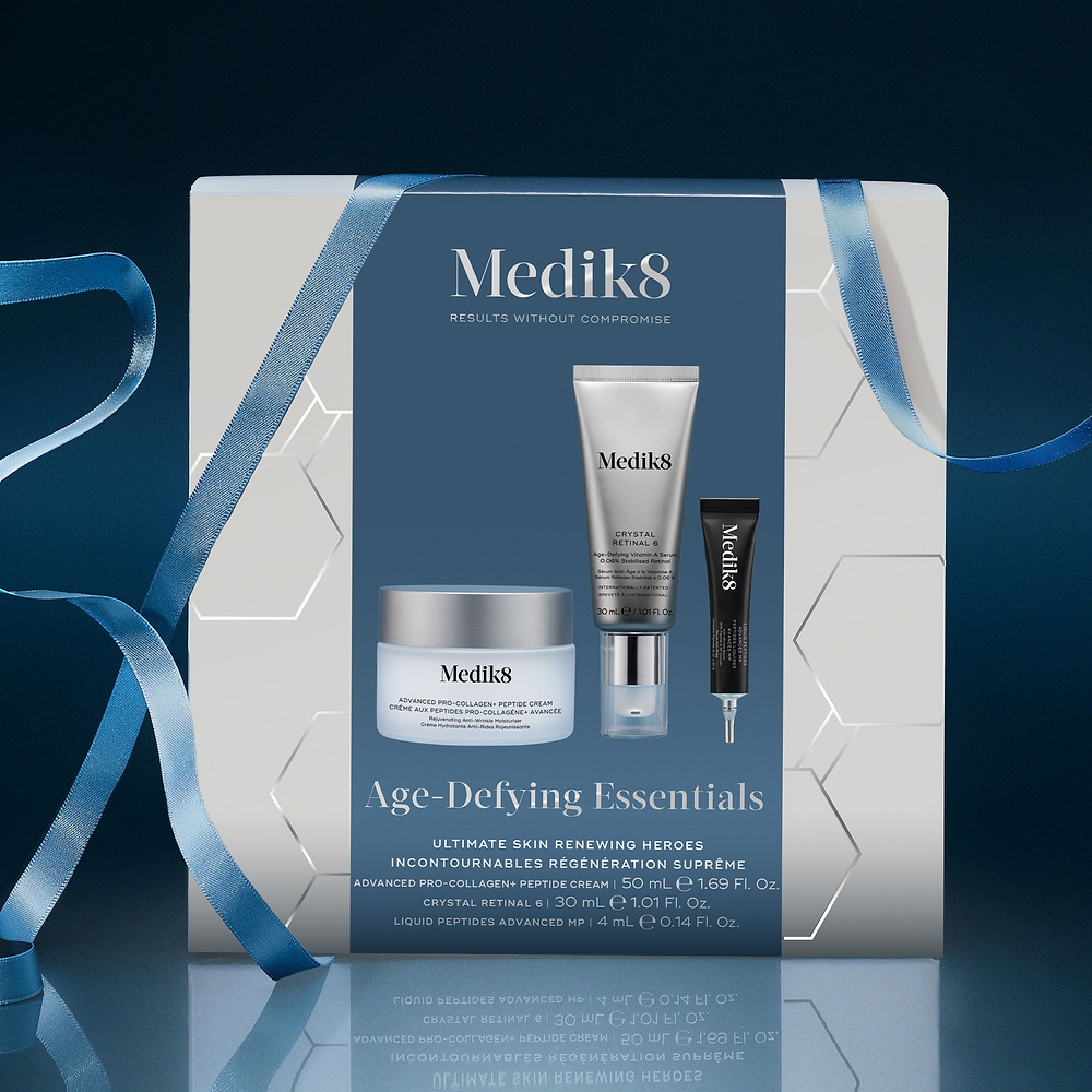 A Medik8 "Age-Defying Essentials" gift box wrapped with a blue ribbon on a reflective surface against a dark blue background. The white box features a dark blue center panel displaying the printed products inside: an Advanced Pro-Collagen+ Peptide Cream jar, a Crystal Retinal 6 tube, and a Liquid Peptides Advanced MP deluxe sample. Text highlights "Ultimate Skin Renewing Heroes."