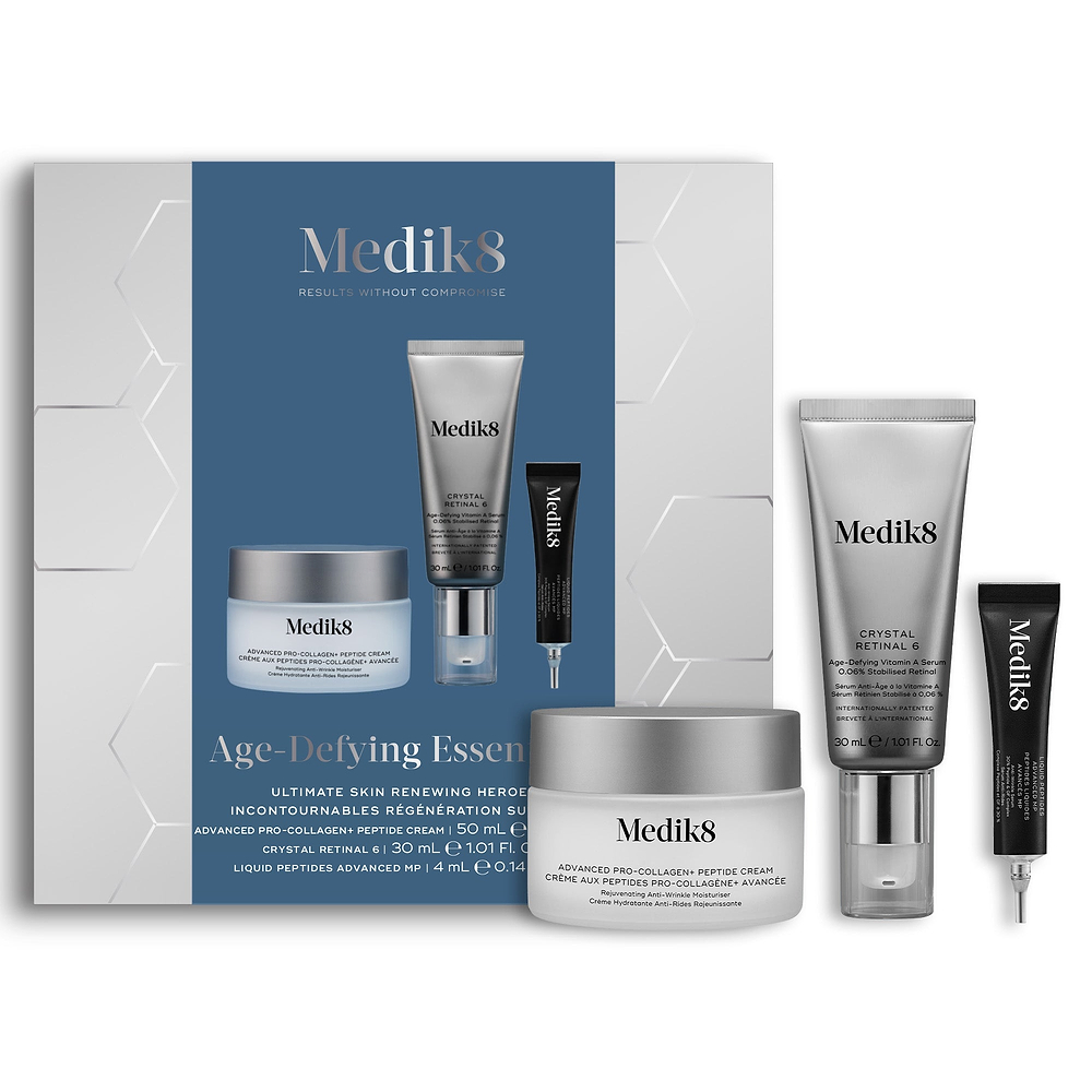 A Medik8 "Age-Defying Essentials" gift box displayed on a white background with a dark blue center and hexagonal graphics, showing the printed products inside. An Advanced Pro-Collagen+ Peptide Cream jar, a Crystal Retinal 6 tube, and a Liquid Peptides Advanced MP deluxe sample stand loose beside the box.
