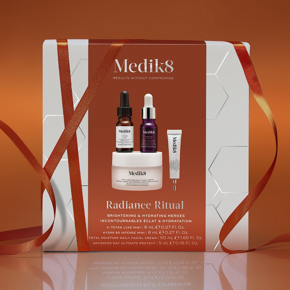 A Medik8 "Radiance Ritual" gift box wrapped with an orange ribbon against a burnt orange background. The white box features a burnt orange centre panel displaying a printed C-Tetra Luxe mini serum bottle, Hydr8 B5 Intense mini serum bottle, Total Moisture Daily Facial Cream jar, and Advanced Day Ultimate Protect deluxe sample tube. Text highlights "Brightening & Hydrating Heroes."