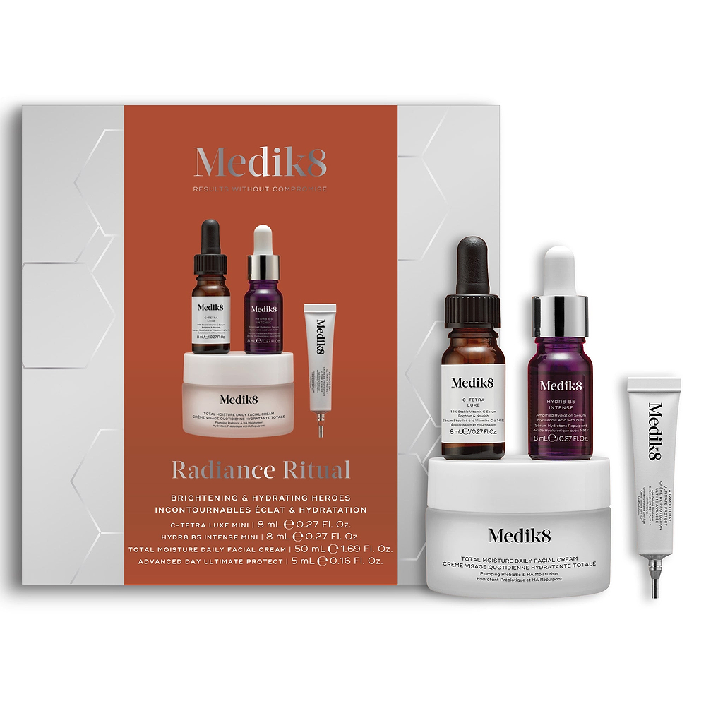 A Medik8 "Radiance Ritual" gift box displayed on a white background with a burnt orange center and hexagonal graphics, showing the printed products inside. A C-Tetra Luxe mini serum bottle, Hydr8 B5 Intense mini serum bottle, Total Moisture Daily Facial Cream jar, and Advanced Day Ultimate Protect deluxe sample tube stand beside the box on a white surface.