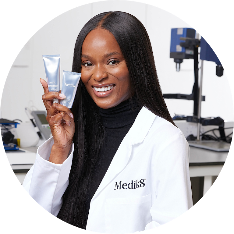 A circular portrait of a smiling skincare professional wearing a branded lab coat, holding two silver Crystal Retinal tubes in a brightly lit laboratory setting.