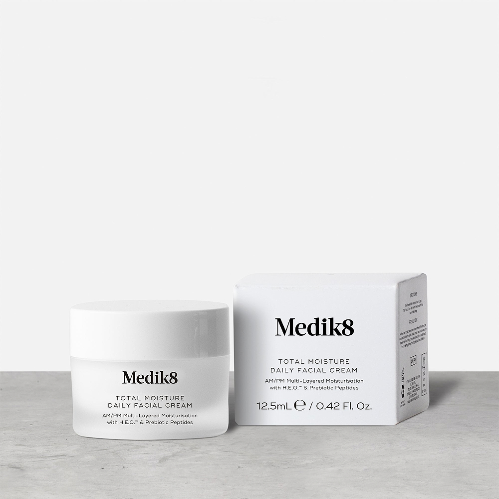 A travel-sized 12.5mL frosted jar of Medik8 Total Moisture Daily Facial Cream standing next to its white cardboard carton. The products are displayed on a textured grey surface against a clean white background. Text on both reads: "AM/PM Multi-Layered Moisturisation with H.E.O.™ & Prebiotic Peptides."
