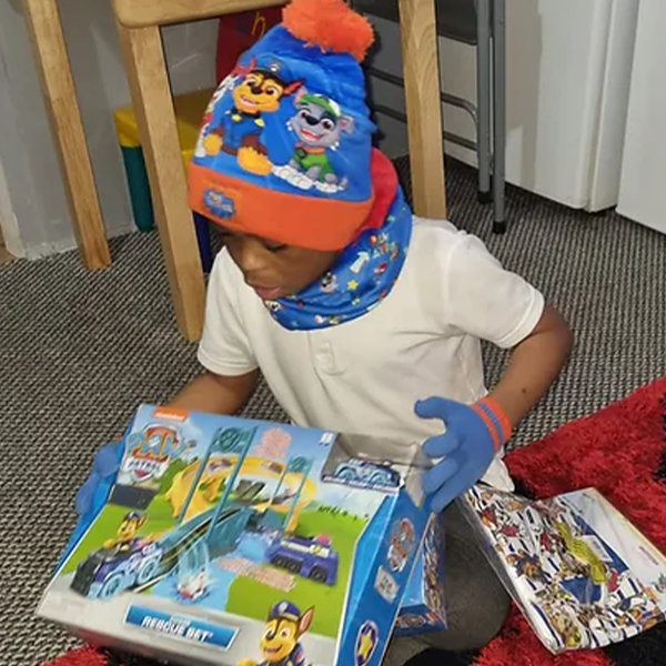 A young child wearing a blue and orange character-themed beanie and matching neck warmer sits on a grey carpeted floor, looking down at an open book. 