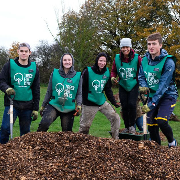 Six volunteers stand together in an outdoor park area, wearing green "Trees for Cities" tabards over their warm clothing. Some hold shovels and stand behind a large mound of wood mulch. 
