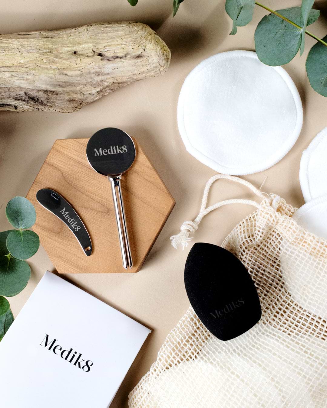 A flat-lay composition of Medik8 eco-accessories on a beige background, including a metal tube squeezer, metal spatula, reusable bamboo cotton pads, a mesh bag, and a black blending sponge.