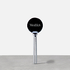 A silver Medik8 metal tube squeezer standing upright on a grey surface. The circular head features the "Medik8" logo, designed to help dispense product efficiently.