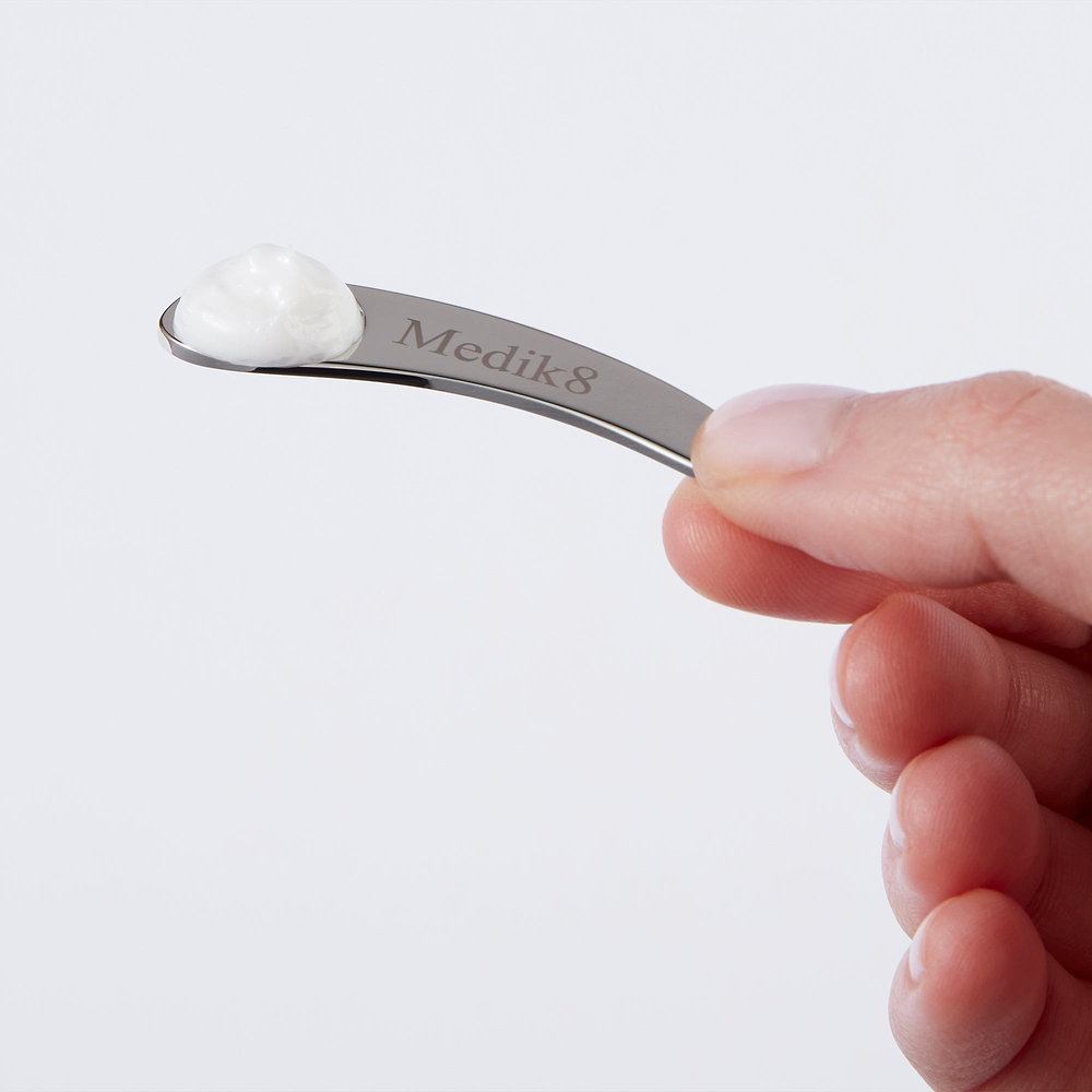 A close-up of a hand holding a Medik8 branded metal spatula with a small dollop of white cream on the tip, demonstrating the correct amount to use.